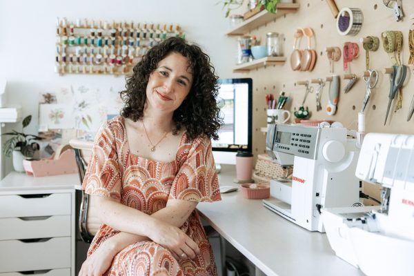 Megan Nielsen sitting at her corner desk with her sewing machines and plywood pegboard wall